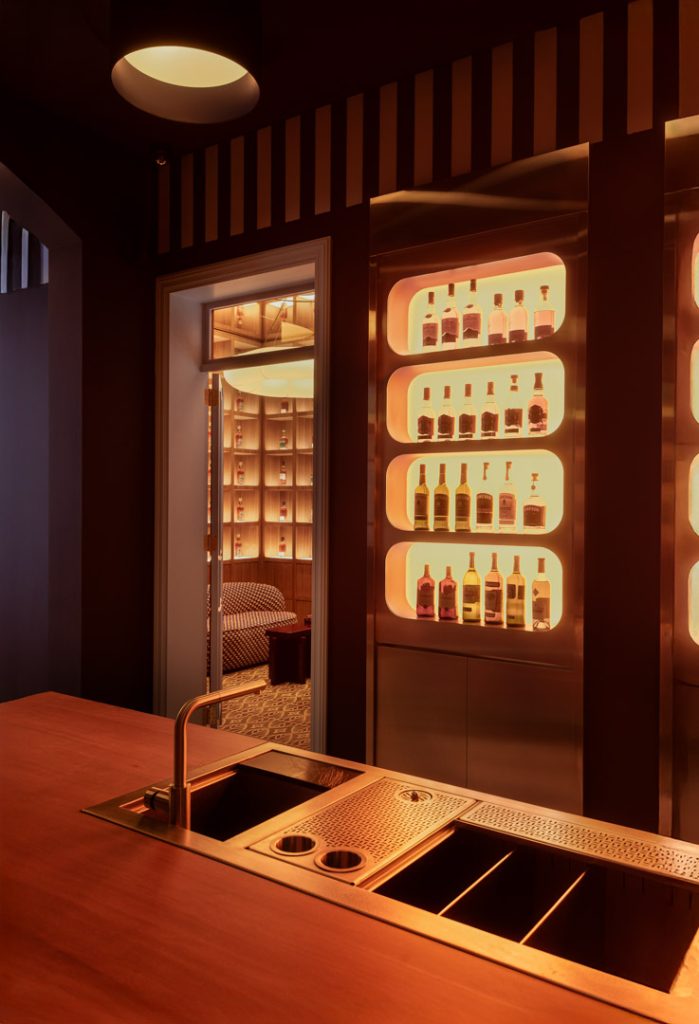 Copper-toned bar interior with backlit rounded bottle shelves and a wooden counter in warm lighting