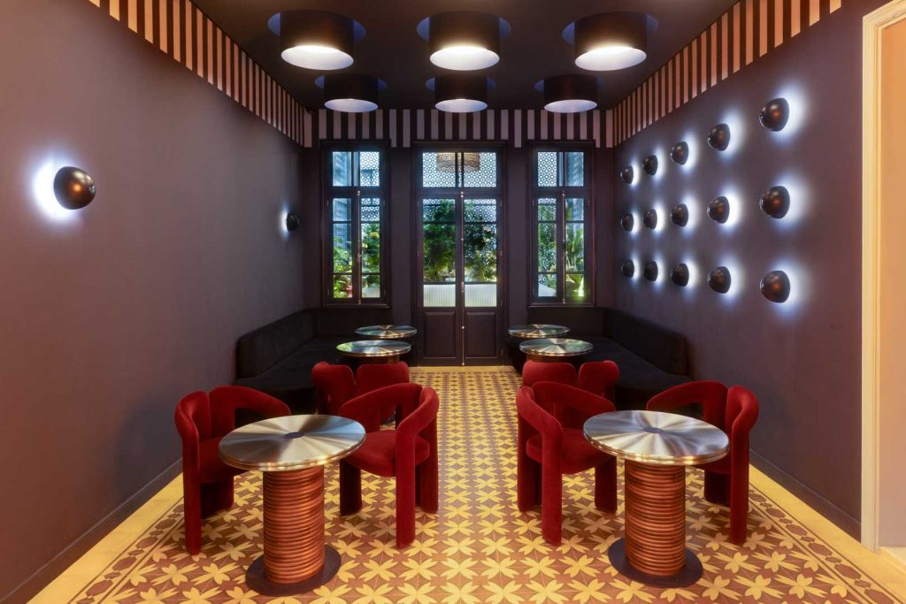 Cozy retro lounge with red curved chairs around shiny metal tables, geometric tiled floor, dark purple walls, and circular wall lights.