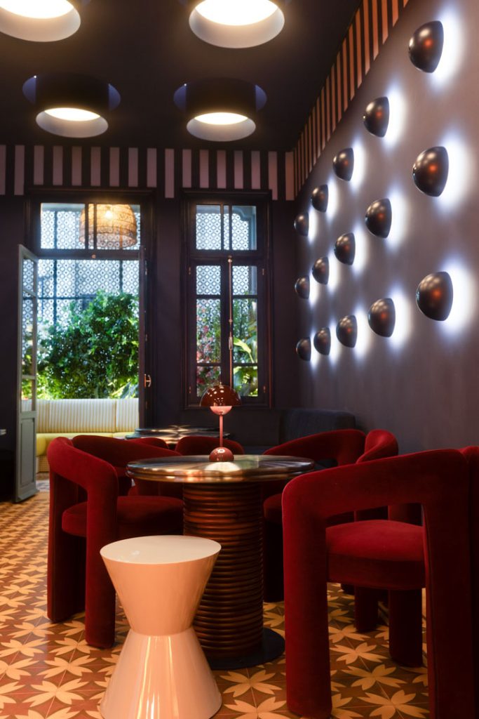 Chic lounge with red velvet chairs around a round ribbed table; patterned tiled floor and purple walls with dome lights, open doorway to greenery outside.
