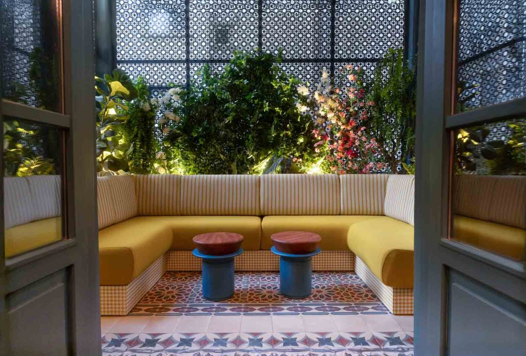 Cozy indoor seating nook with yellow cushioned benches, two small round-topped tables, and a lush green plant wall behind patterned panels.