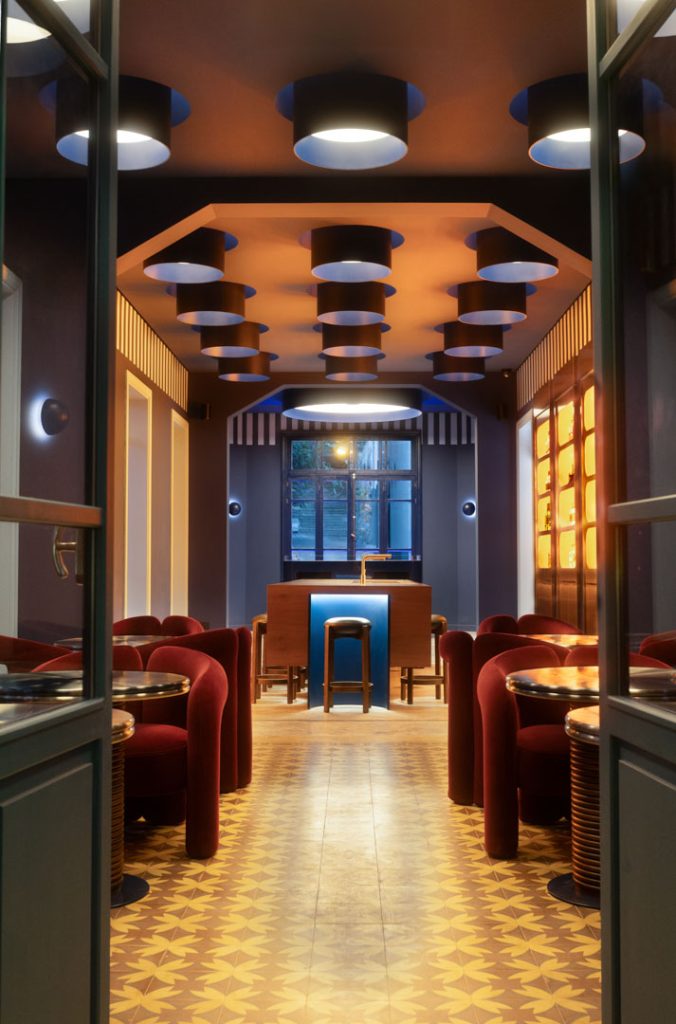 Café interior with red curved seating, patterned tile floor, and a blue-lit bar at the far end under circular ceiling lights.