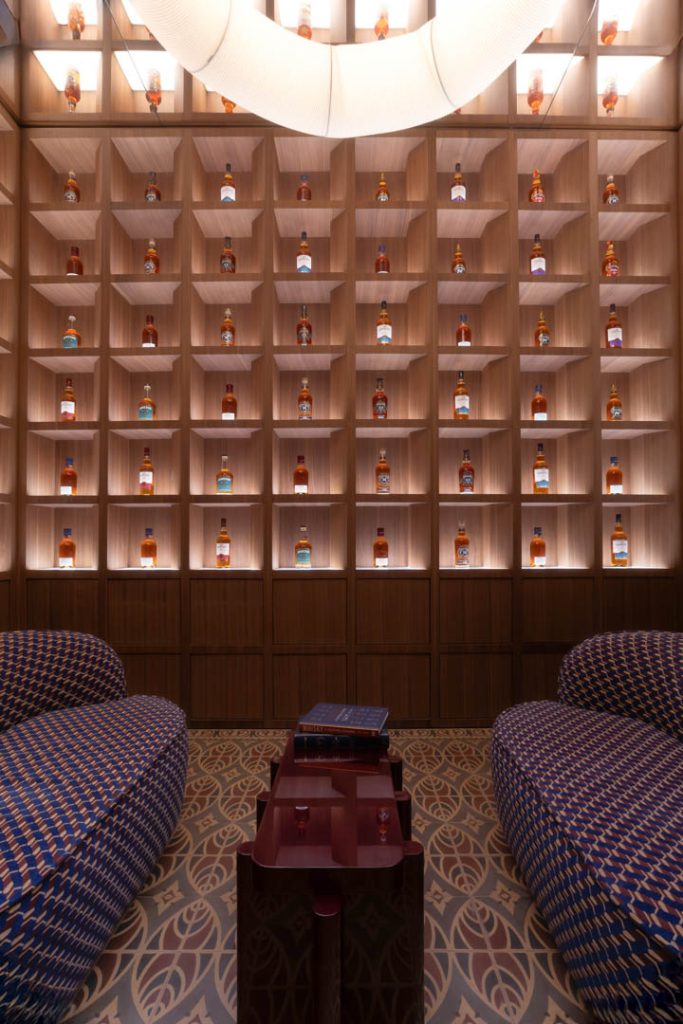Lounge with a wall of backlit bottle alcoves behind seating, two blue patterned sofas, and a wooden coffee table with books.