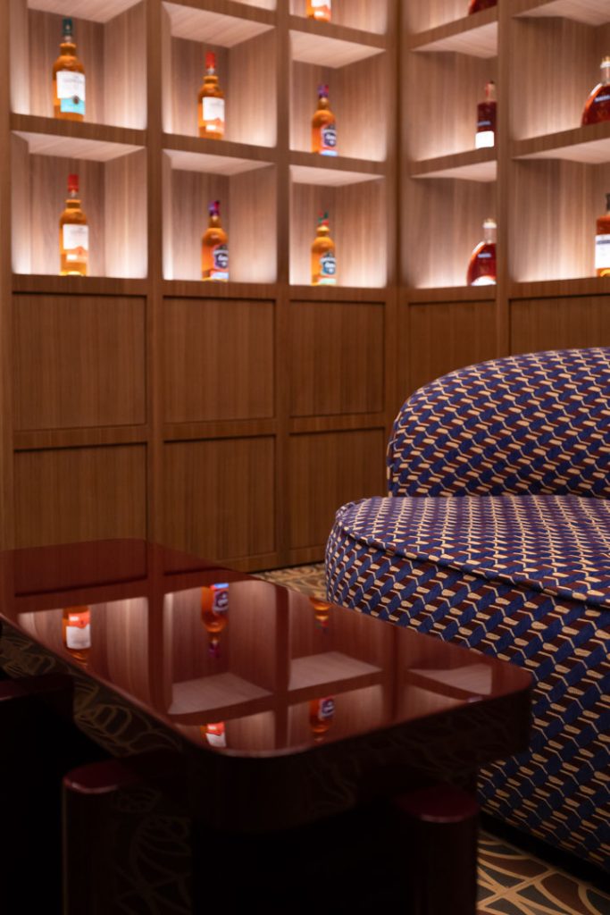Lounge area with backlit bottles on wooden shelves, a blue patterned sofa, and a glossy coffee table reflection.
