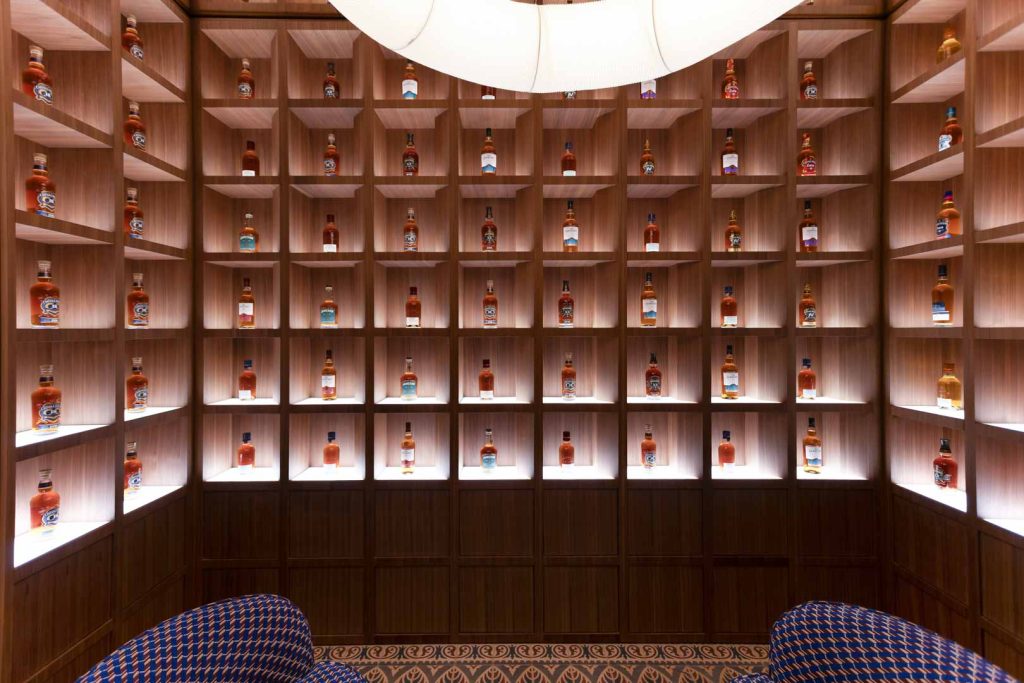 Wall of wooden cubby shelves filled with whiskey bottles, warmly lit tasting room ambiance.