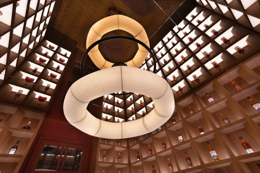 Luxurious bar interior with tall wooden shelves filled with bottles on both sides and a large circular illuminated sculpture hanging from the ceiling