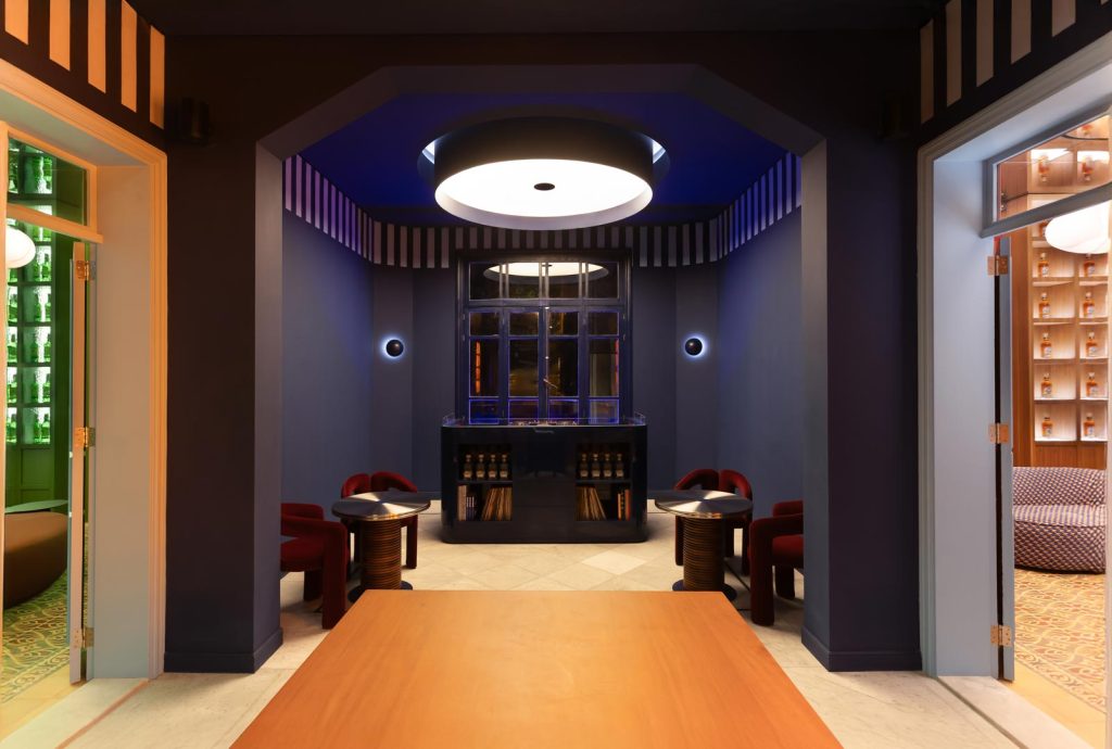 Modern, dimly lit lounge area with blue walls, a central black bar, and red upholstered chairs around small round tables under a large circular ceiling light.