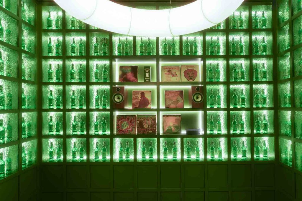 Green-lit room with glass shelves filled with bottles and a central display of vinyl records and speakers on illuminated cubbies