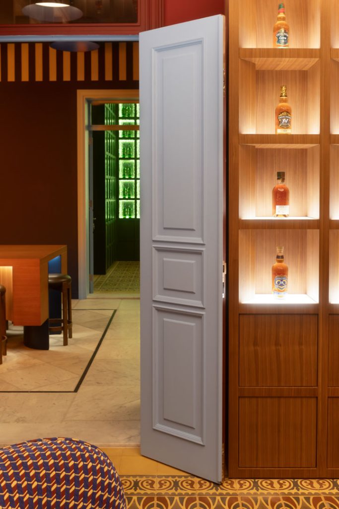 Open light-blue paneled door reveals a hallway with green glass blocks at the far end; a wooden liquor cabinet with lit shelves sits to the right.