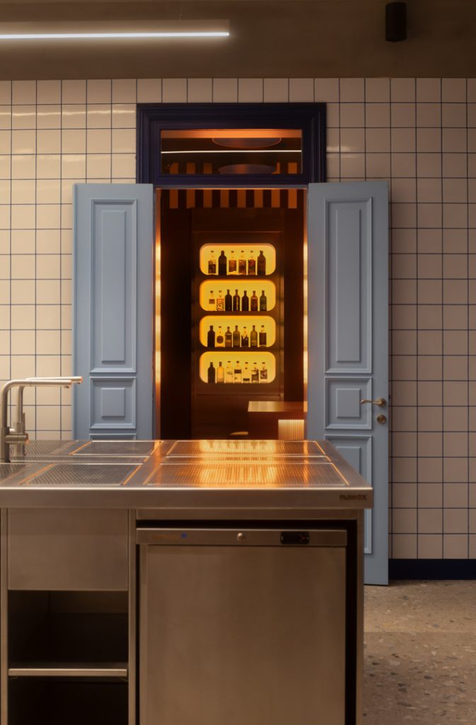 Blue double doors open to a warmly lit liquor display behind a stainless steel kitchen island with tiled walls in a modern setup.