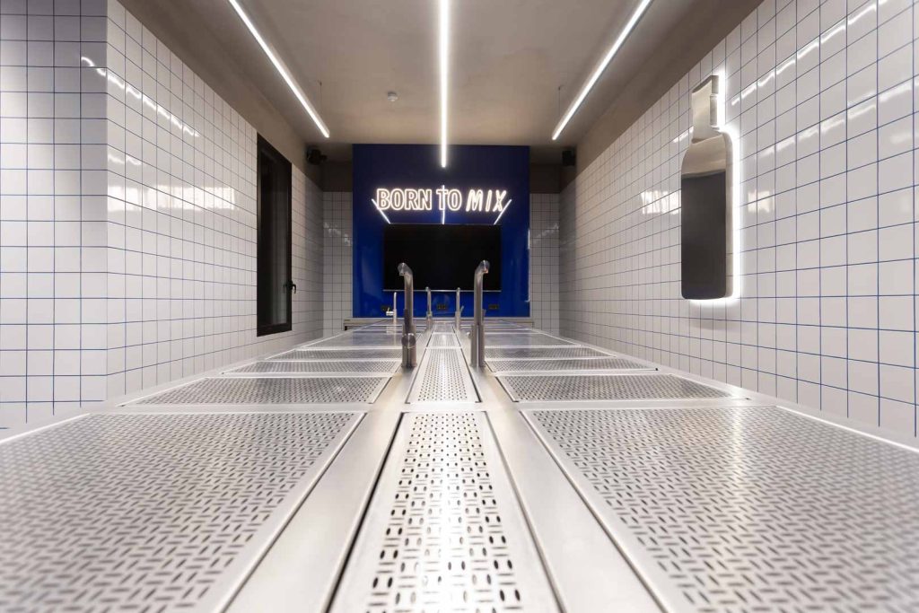 Industrial hallway with perforated stainless benches and white tiled walls leading to a blue wall featuring a neon 'BORN TO MIX' sign at the end.