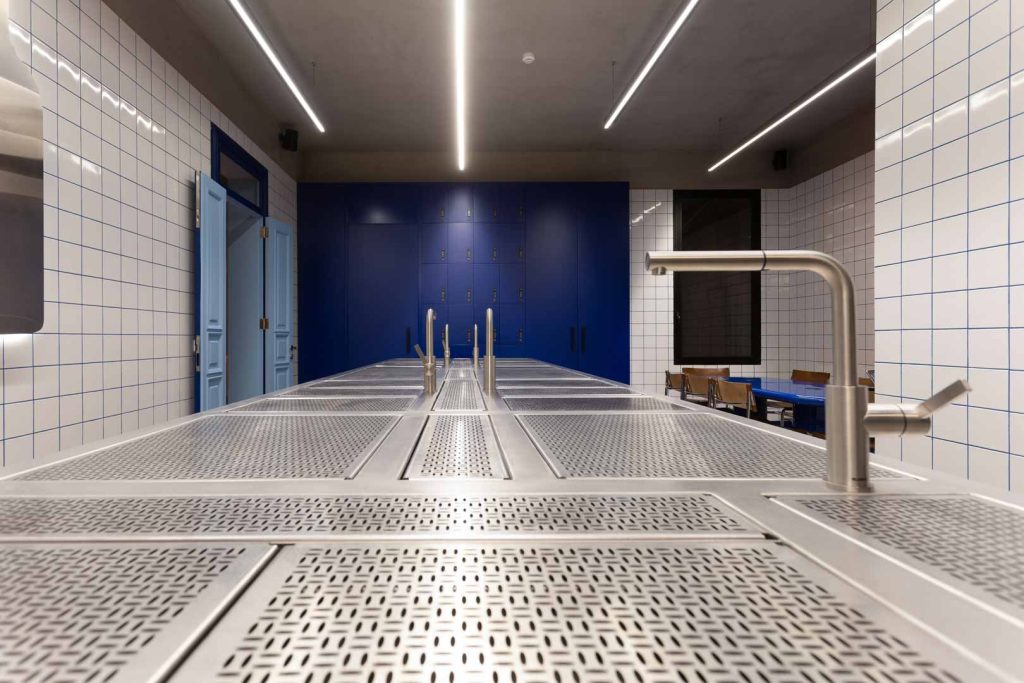 Industrial locker room or prep area with blue lockers along white tiled walls and a long perforated stainless workbench with sinks.