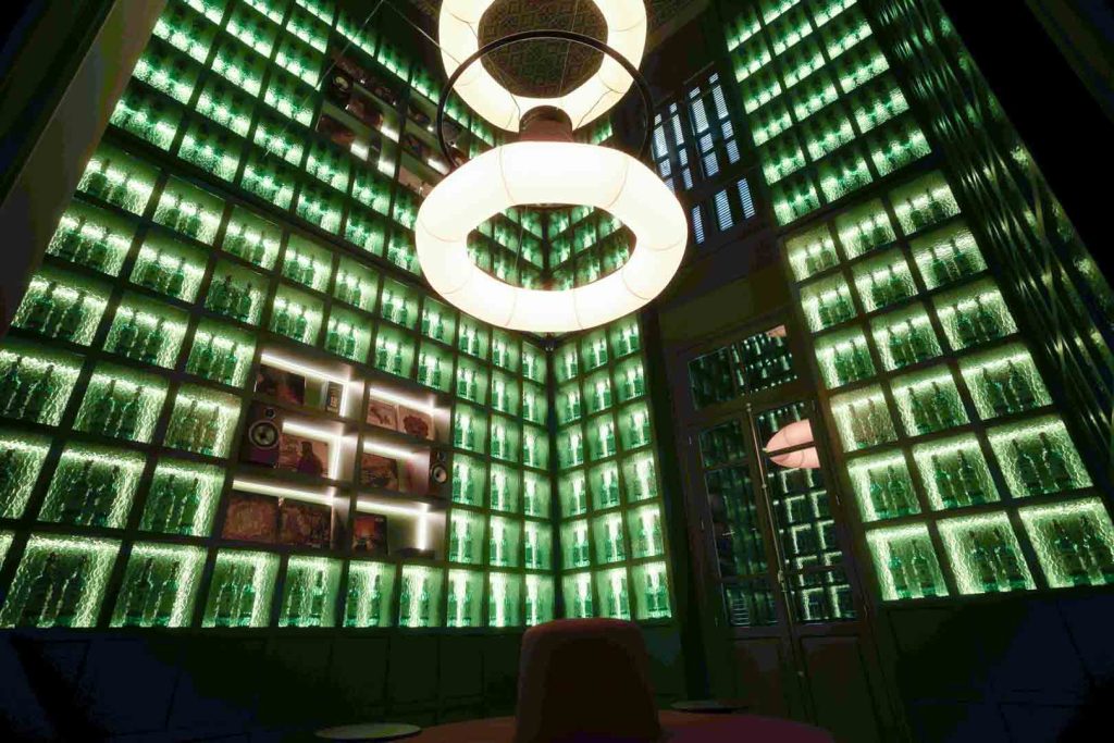 Luxurious bar interior with green-lit wall of shelves filled with bottles and a large circular ceiling light.