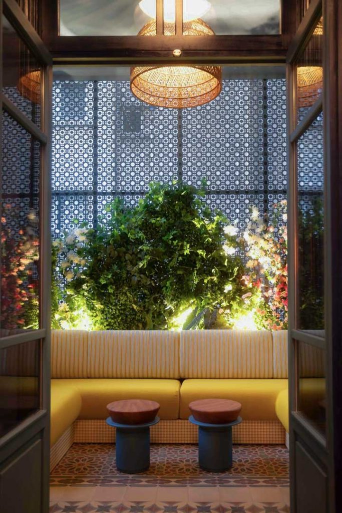 Cozy indoor seating area with yellow striped booth seating facing a large leafy green plant; two blue pedestal tables with wooden tops nearby.