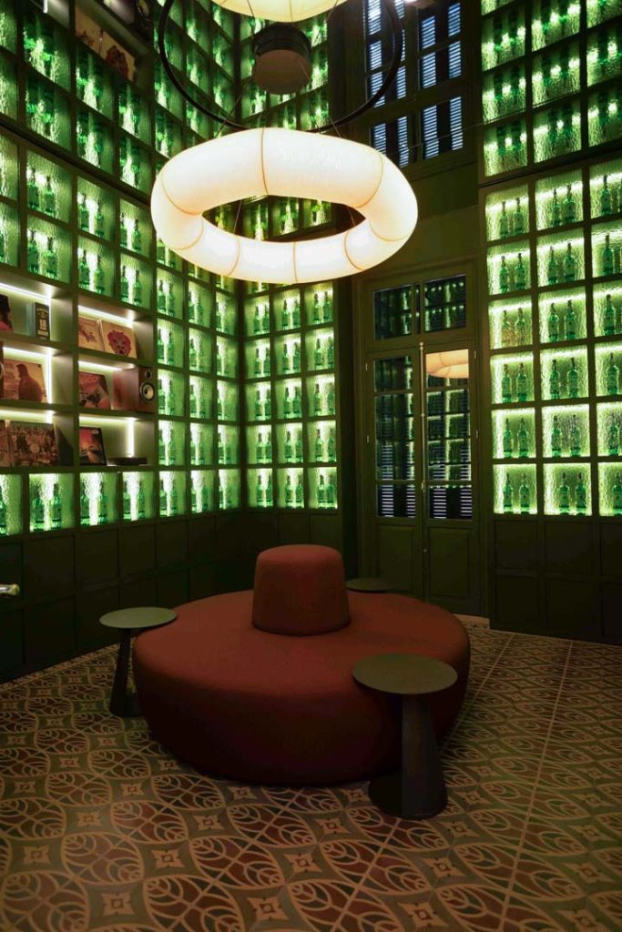 Luxe lounge with green backlit bottle shelves lining the walls, a circular red sofa in the center and small side tables.