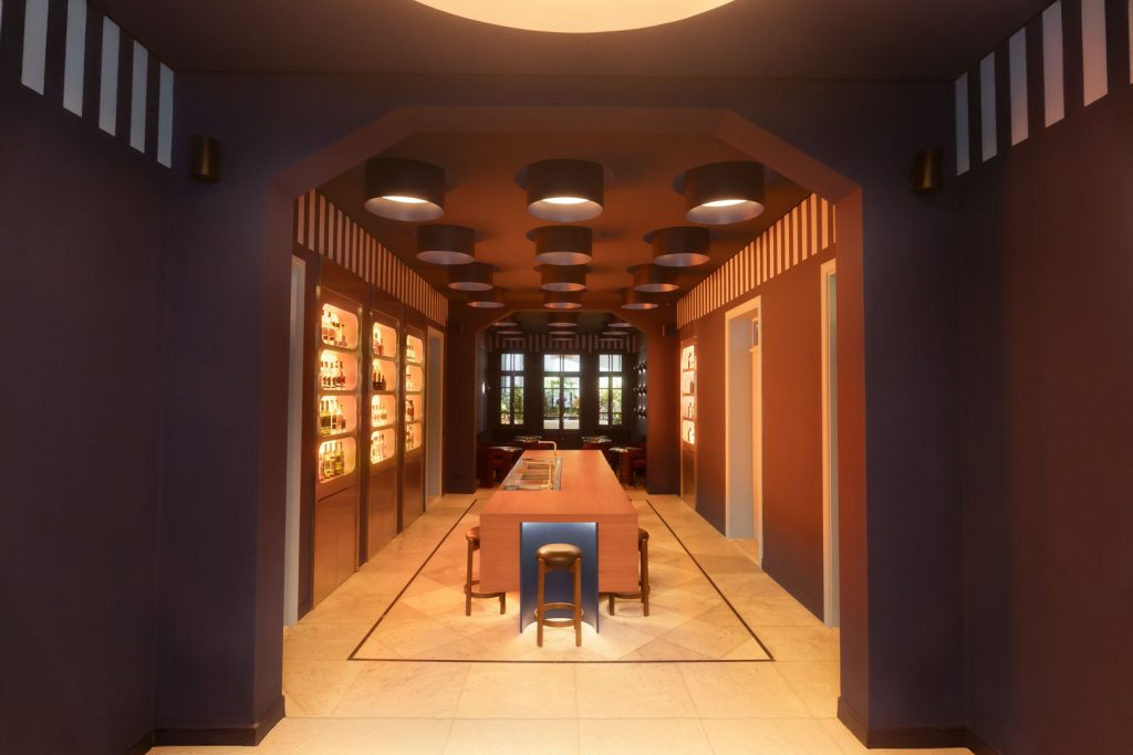 Interior of a modern wine-tasting room with a central wooden island, round pendant lights, and bottle displays along the left wall.