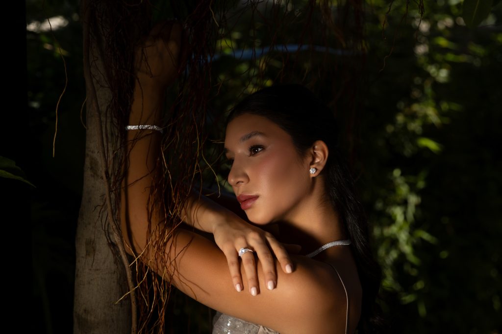 Portrait of a woman wearing diamond earrings and a necklace, leaning against a tree with vines and soft sunlight on her face.