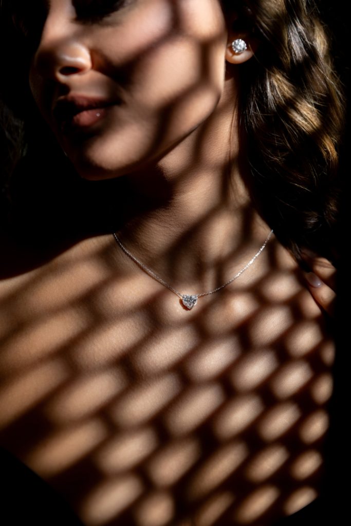 Woman's neck and lower face in shadow, diamond heart pendant necklace visible with matching diamond stud earrings, lattice shadow pattern across skin