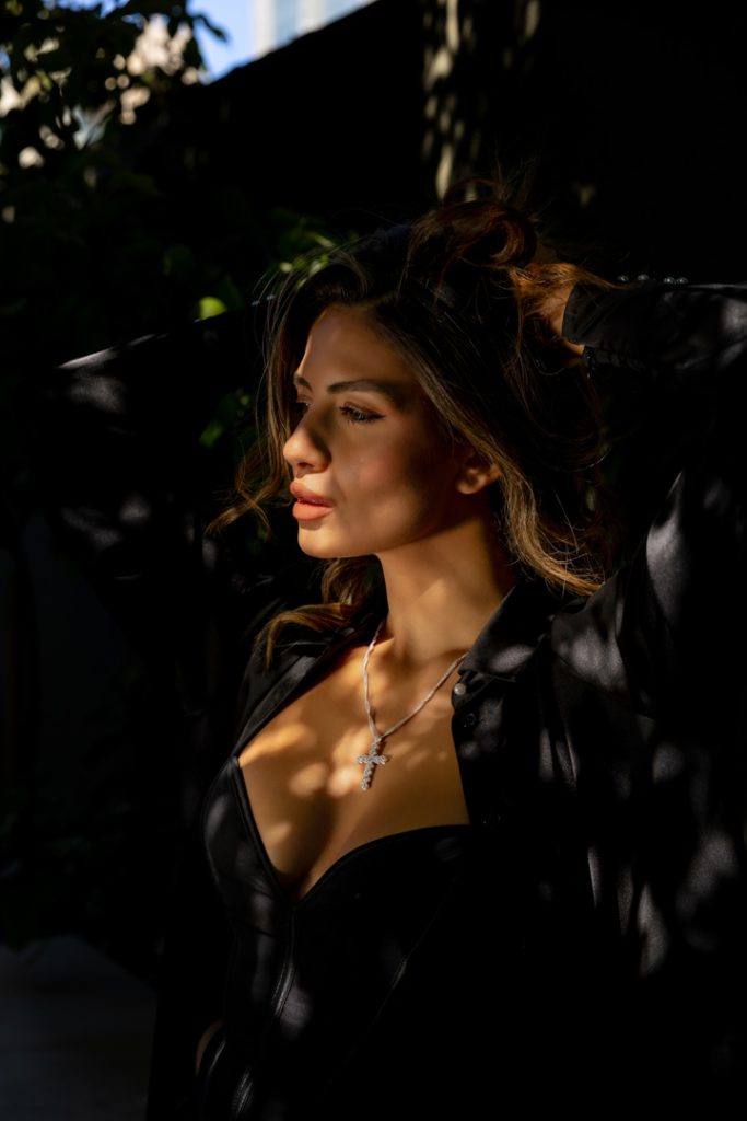 Portrait of a woman in a black outfit with a cross pendant necklace, illuminated by dappled sunlight outdoors, arms raised behind her head