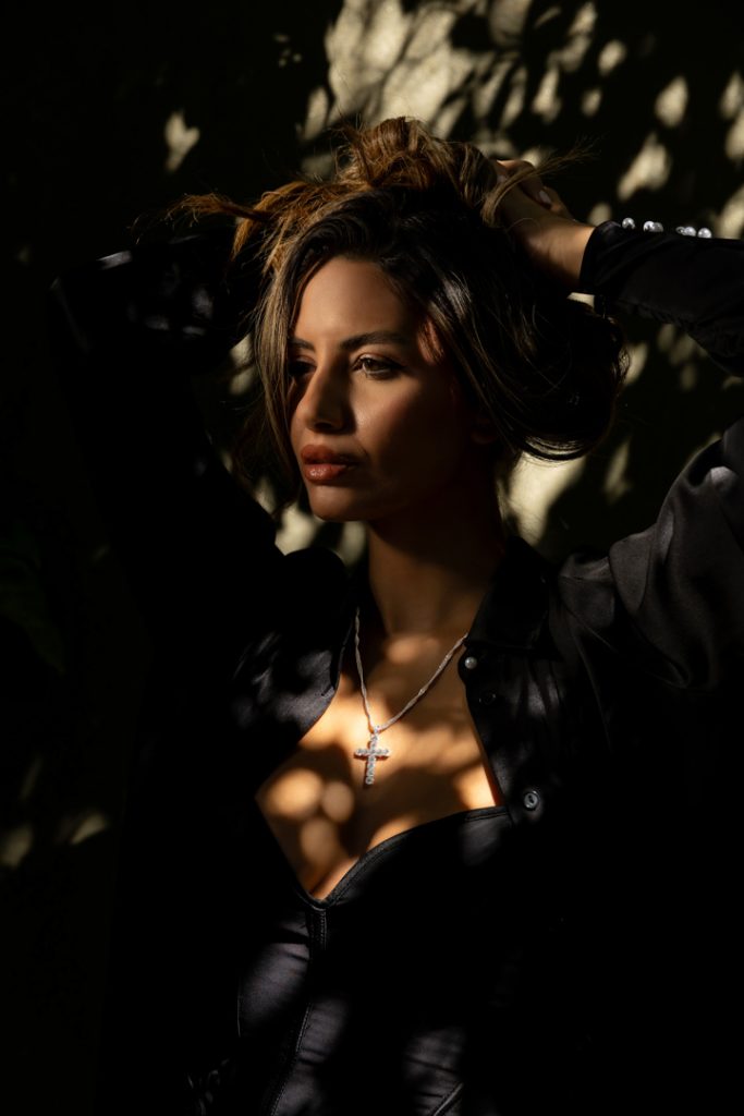 Woman in a black outfit poses with hands in her hair as sunlight and shadows fall on her face, wearing a cross necklace.