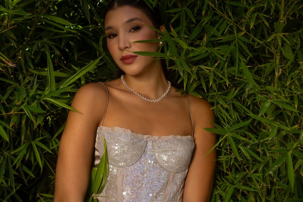 Woman posing among dense bamboo, wearing a sparkly silver dress and a diamond-like necklace with soft makeup and red lips.