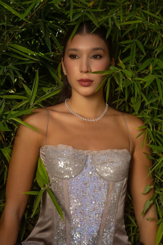 Woman in a shimmering silver evening gown framed by dense bamboo leaves, wearing a diamond necklace.
