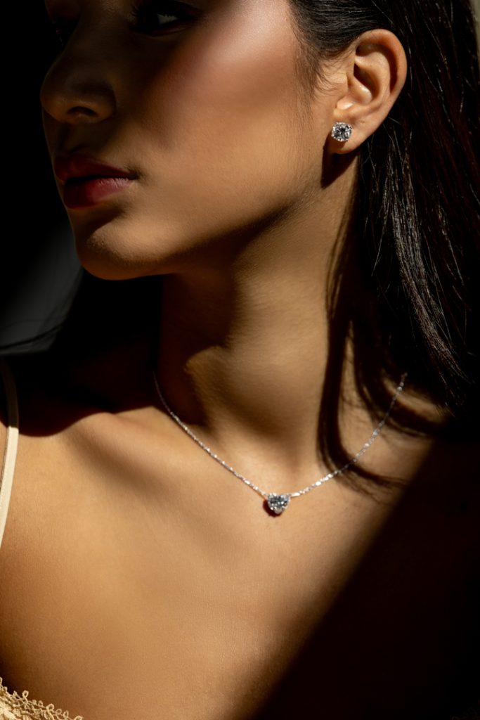 Close-up of a woman wearing a diamond heart pendant necklace and diamond stud earrings, highlighted by warm light.